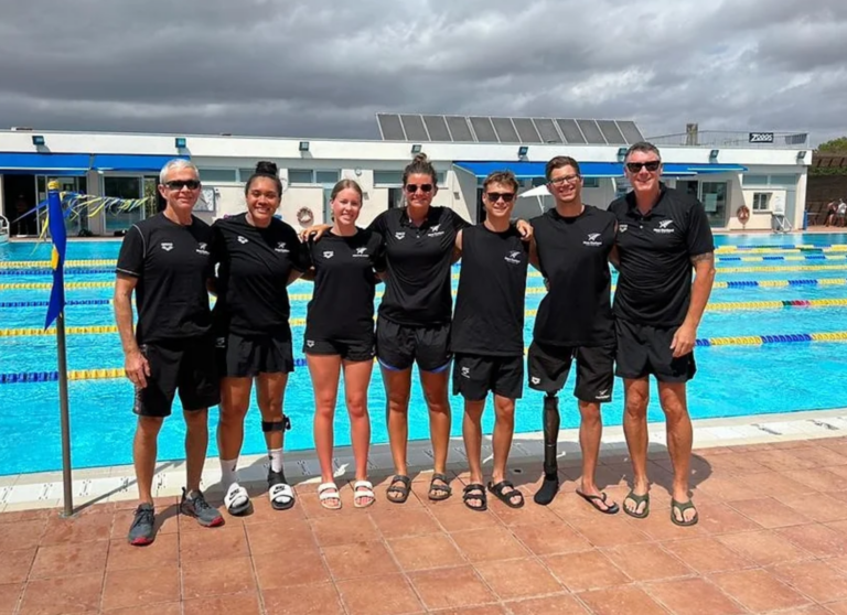 Most of the swim team and support staff stand in a line in front of an outdoor pool in uniform