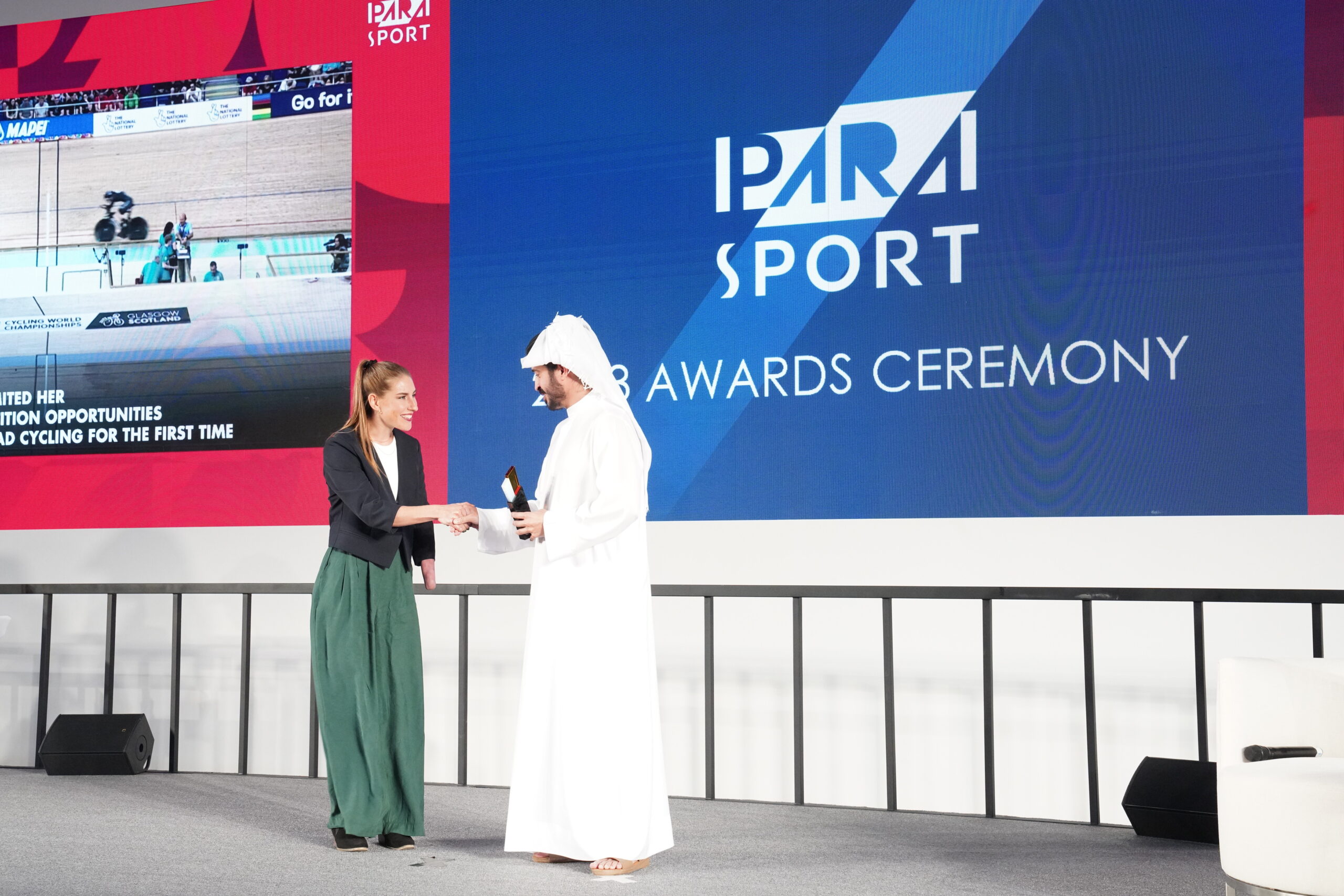 On a stage, with the words "Para sport awards ceremony" behind, a slender fair haired woman shakes hands with a man in a white arab robe and headscarf