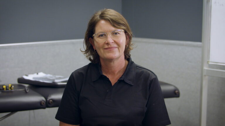 Becs, a caucasian woman wearing a black shirt and glasses, smiles at the camera. There's a physio bed in the background