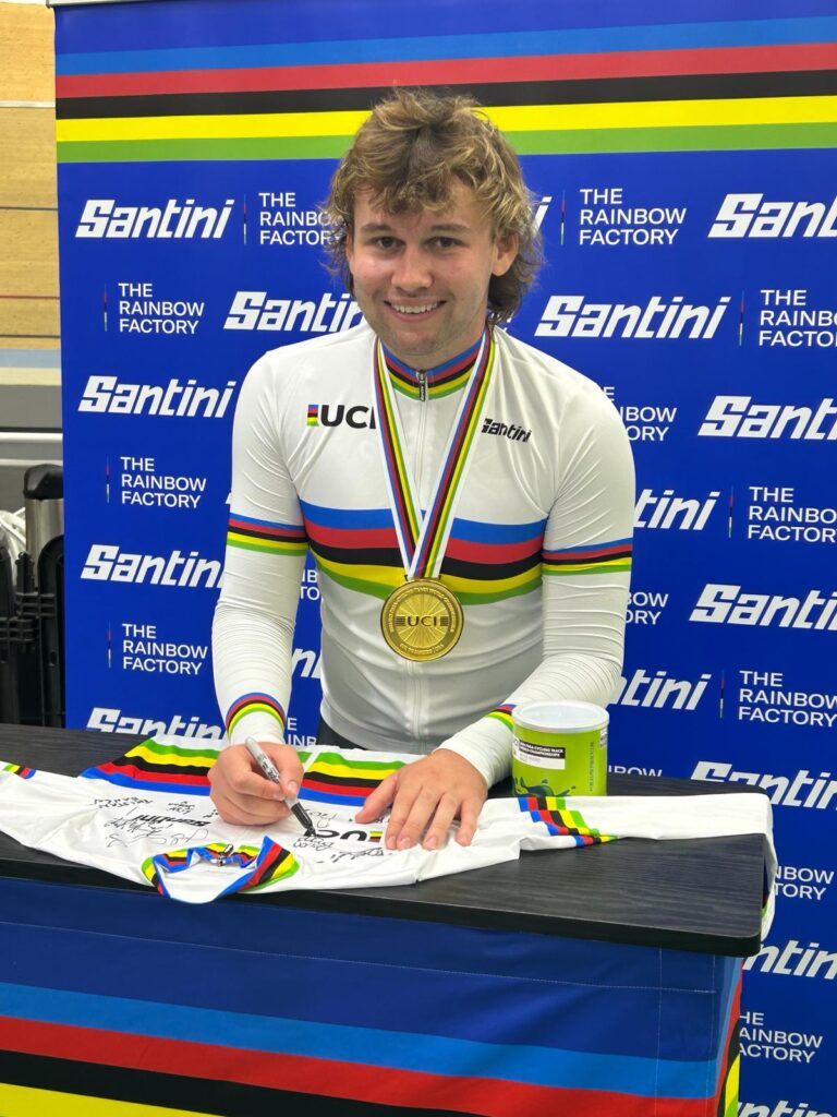 Devon wears the rainbow jersey and his gold medal as he signs another rainbow jersey, smiling.