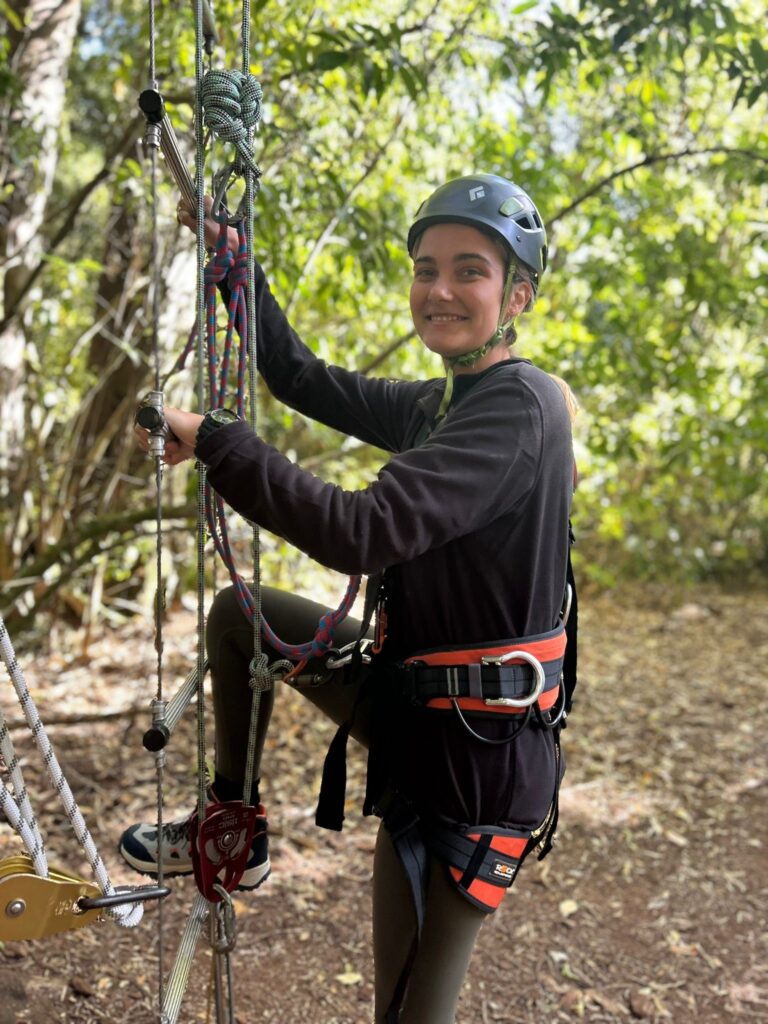 Georgia smiles as she puts one foot up on a rope ladder in the forest