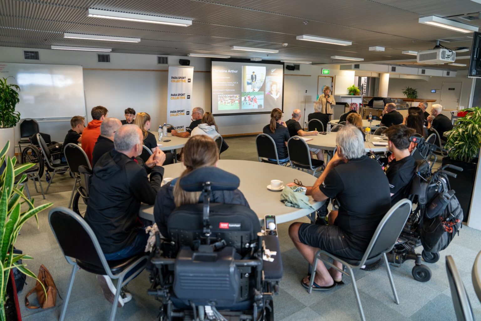 A group of participants sits and watches a presenter talk to a slide